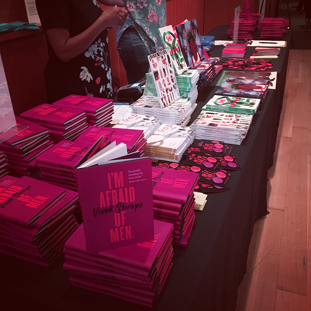 A photo of the vendor table at the launch of I'm Afraid of Men, covered in many copies of Vivek Shraya's books, records, and children's books. There are also lots of I'm Afraid of Men buttons and bookmarks.