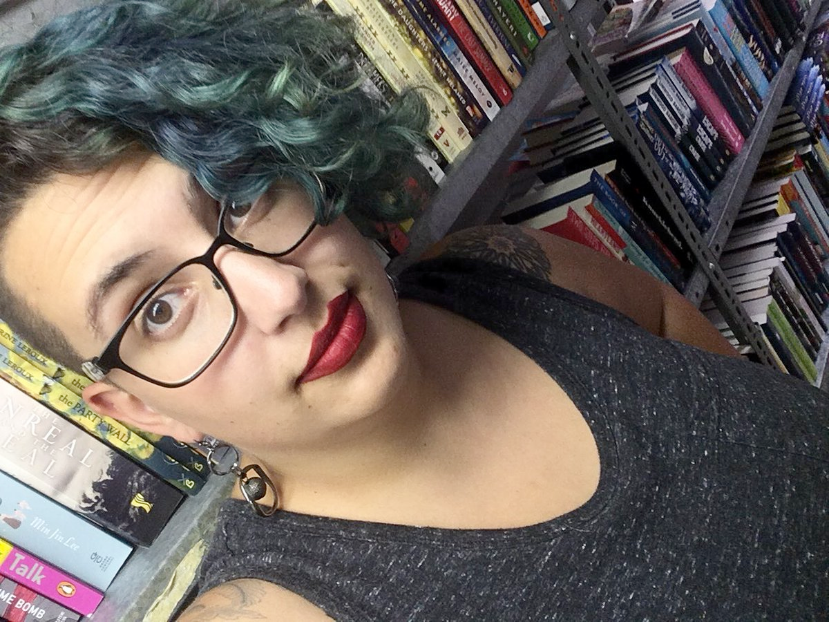 This image is a selfie of me with teal hair and red lipstick. It's taken in the basement of the book shop where I work, so there are shelves of overstock books in the background.