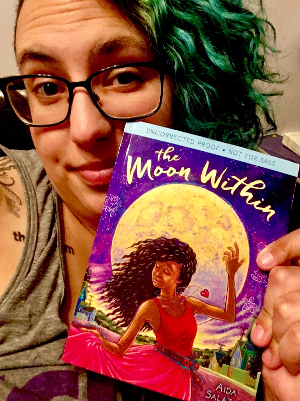 A selfie of me, with green hair, looking sleepy, and holding an ARC of The Moon Within, by Aida Salazar.