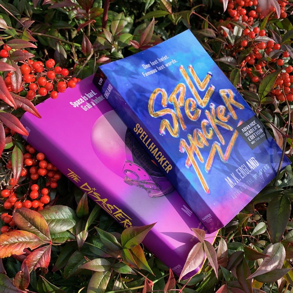 Two books laying in some festive foliage. Underneath, a hardback of The Disasters, a space helmet on a pink background. On top, an ARC of Spellhacker, a purple galaxy print cover with sparkly gold text.