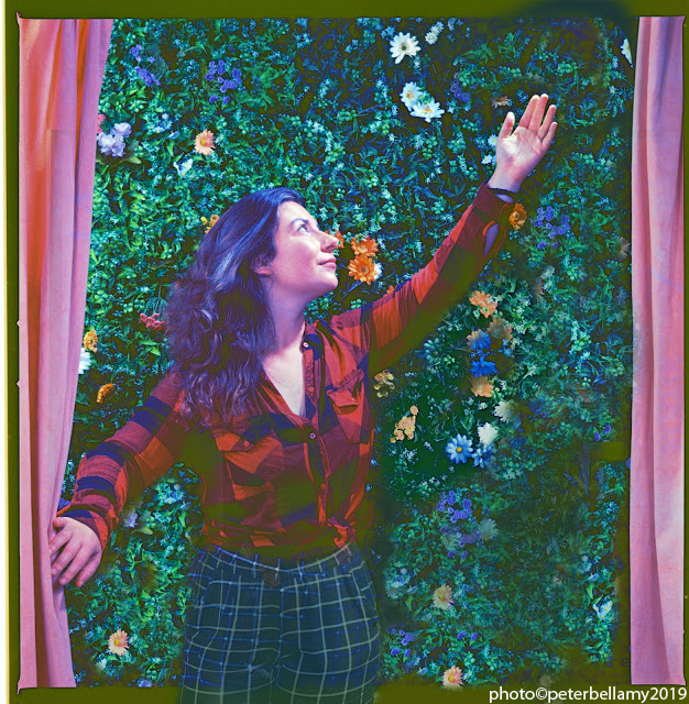 A stylized photo of Chana Porter, looking upward, arm reaching up. Theatre curtains frame the image. The background is foliage with flowers.