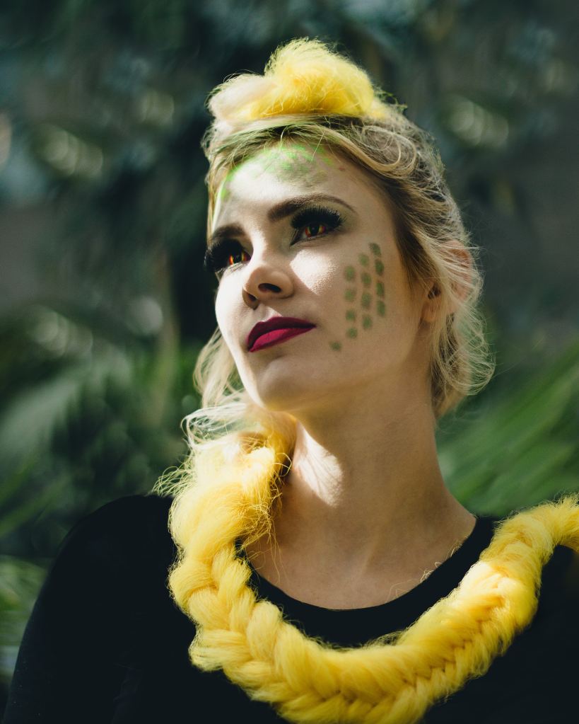 An image of a white person with red lipstick, snake eye contacts, and green scales airbrushed onto their cheek. Their hair is bright yellow and long, wrapped around their neck in a braid.