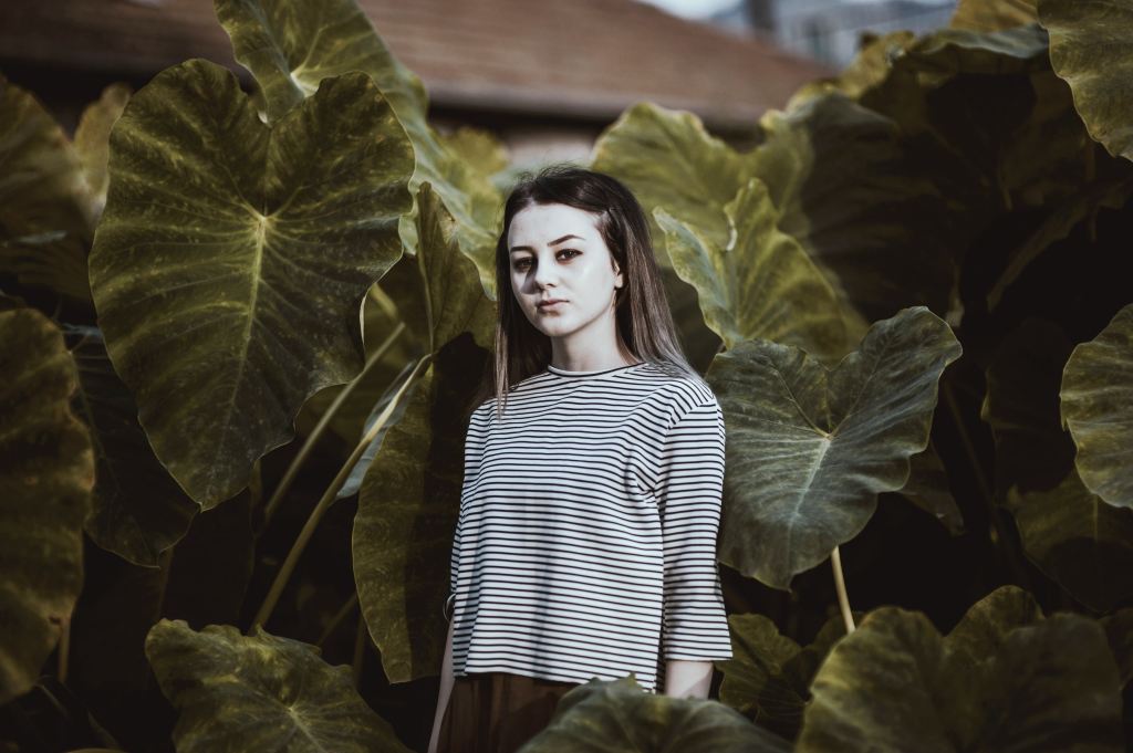 A pale, slightly monochromatic feminine figure stands against a backdrop of very large foliage.