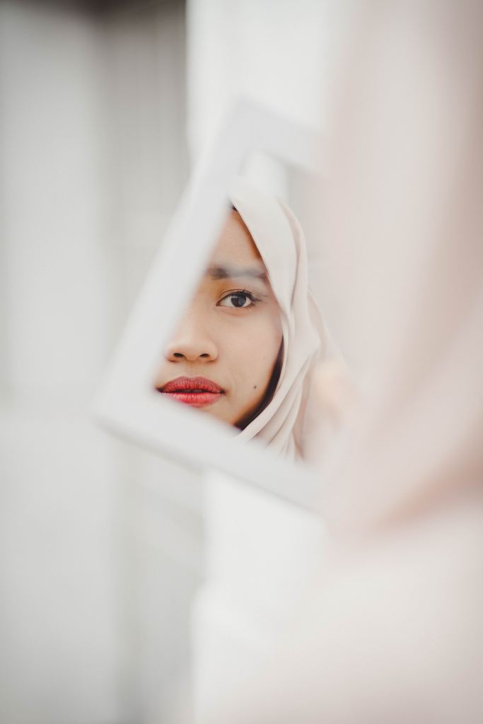 A person's face, partially obscured, wearing a headscarf and reflected in a mirror.