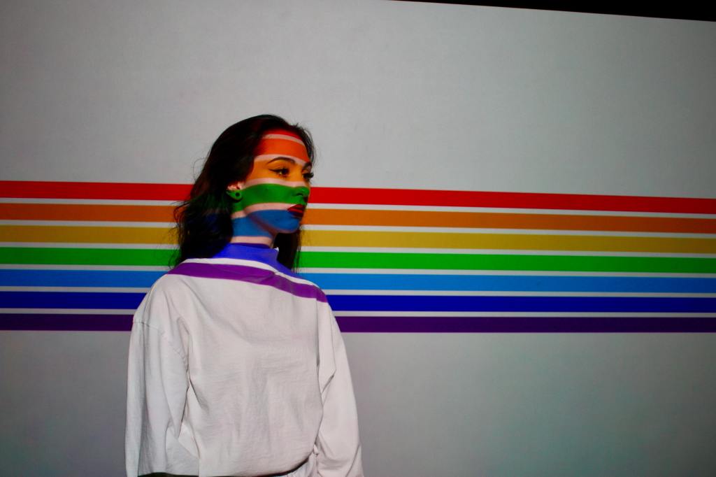 A person standing on a white background with rainbow stripes. The stripes cross their face as well.