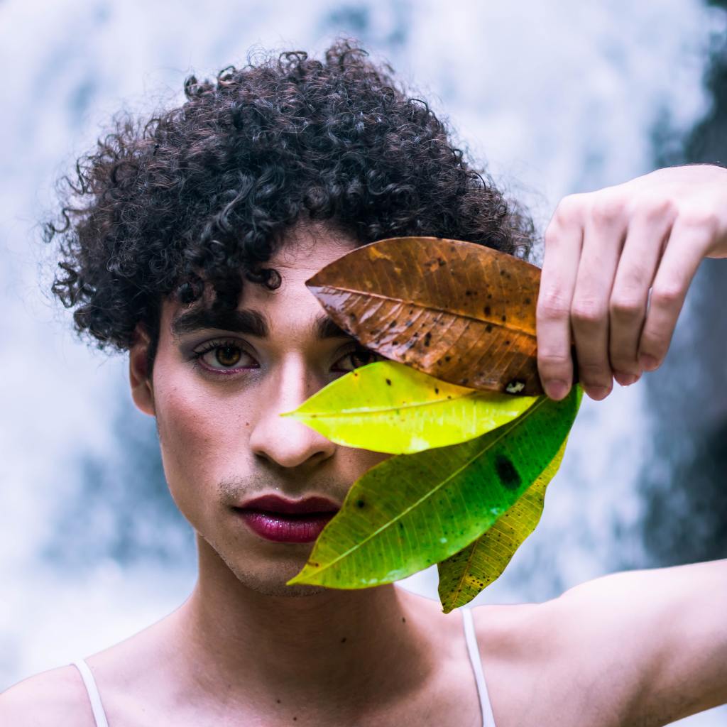A photo of a person wearing sparkly lipstick, with curly hair. They are holding four brightly-coloured leaves over one of their eyes.