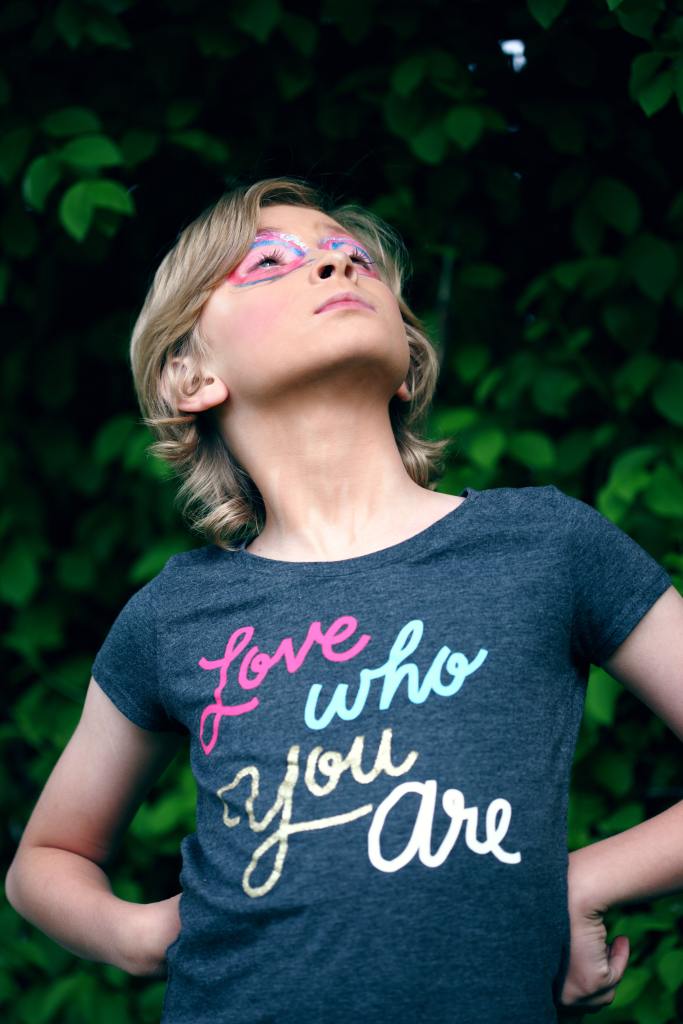 A young person with their hands on their hips, looking upward. They have elaborate eye makeup on, and a t-shirt that says, Love you who are.