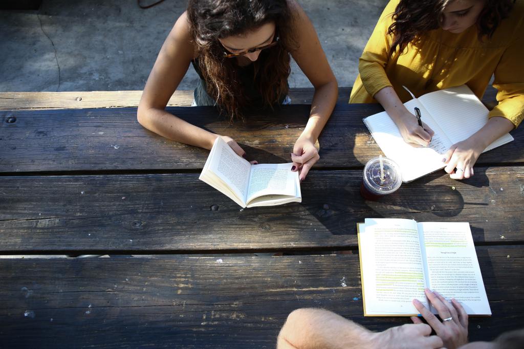 A group of people sitting at a wooden table with notebooks, a plastic coffee cup, and a book with highlighting inside. They look like friends collaborating on a project.
