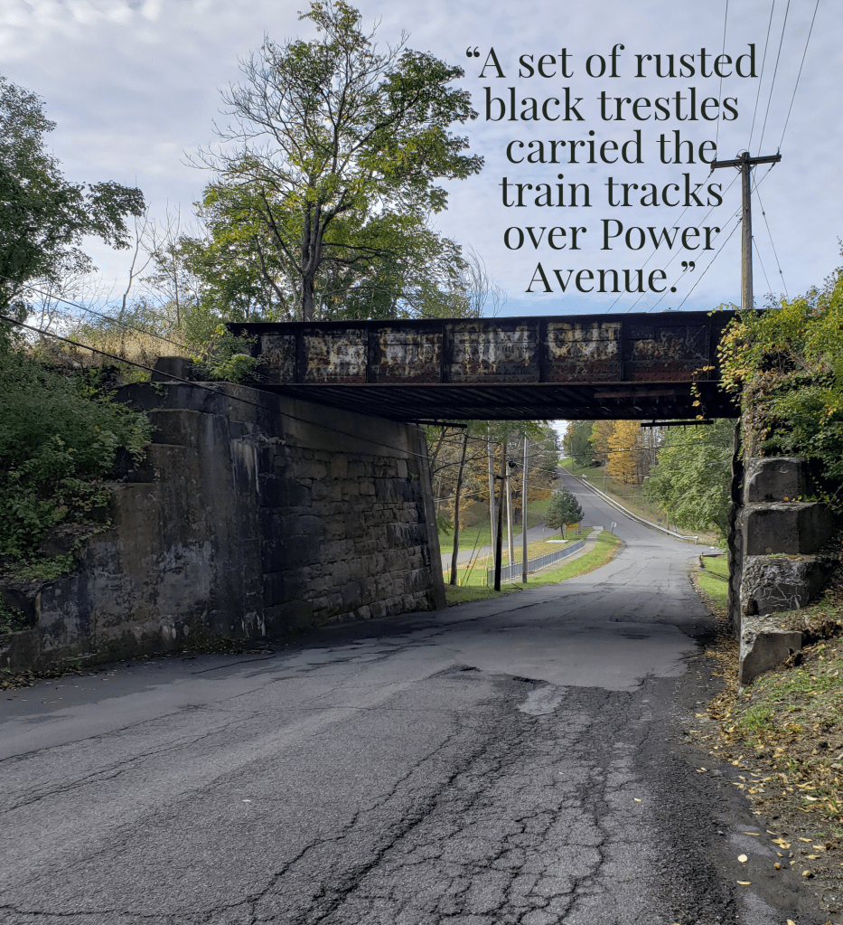 A bridge covered in faded graffiti over a worn concrete road. Text reads, A set of rusted black trestles carried the train tracks over Power Avenue.