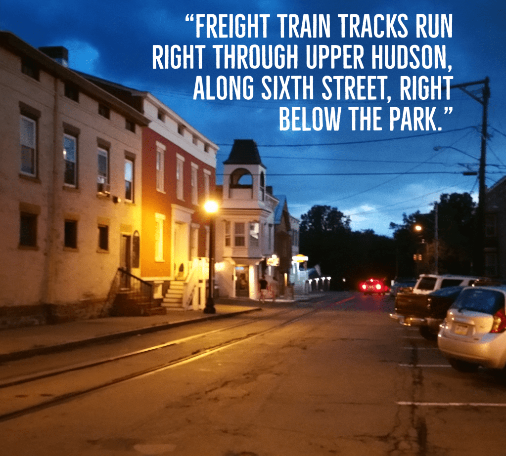 Twilight, a street with train tracks embedded in it, lined on one side with run-down buildings, and on the other side with parked cars. Text reads, Freight train tracks run right through upper Hudson, along sixth street, right below the park.